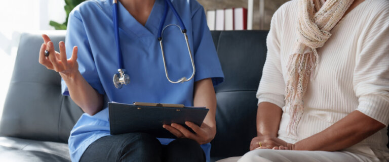 The image shows a healthcare professional in blue scrubs with a stethoscope around their neck, holding a clipboard while speaking with a patient. The patient is seated beside them, wearing a light-colored outfit and scarf, suggesting a medical consultation or discussion.
