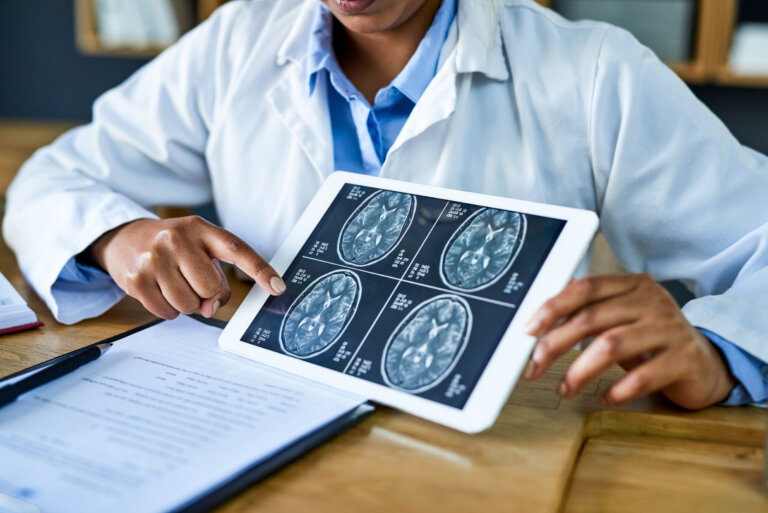 A doctor in a white coat is pointing at brain scan images displayed on a tablet, with medical documents on the desk.