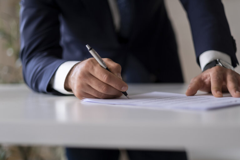 A person in a suit is signing a document on a desk with a pen, focusing on the act of writing.