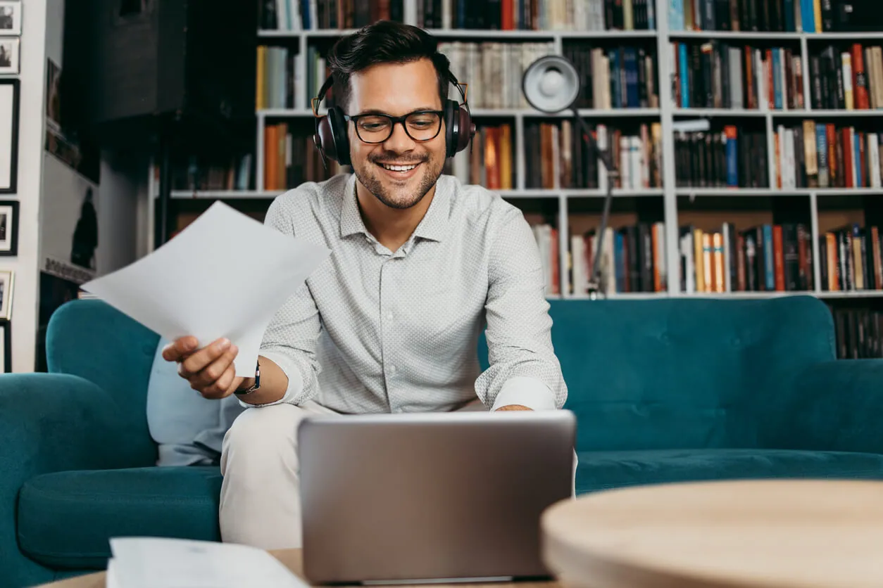 man on green couch wearing head phones going through papers and using a laptop