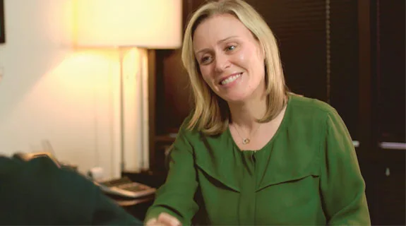 A smiling woman in a green blouse shakes hands with someone in an office setting.